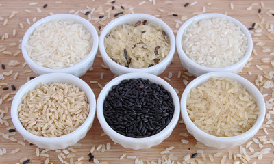 Six varieties of uncooked rice in white bowls on wooden board
