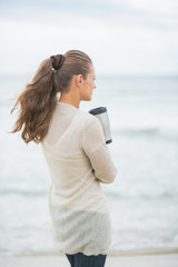 Young woman standing on cold beach with cup of hot beverage