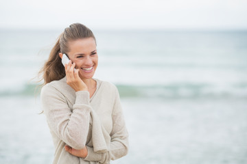 Fototapeta na wymiar Smiling young woman talking cell phone on cold beach