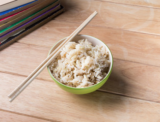 Rice a bowl with chopsticks and a book rests on a wooden desk.