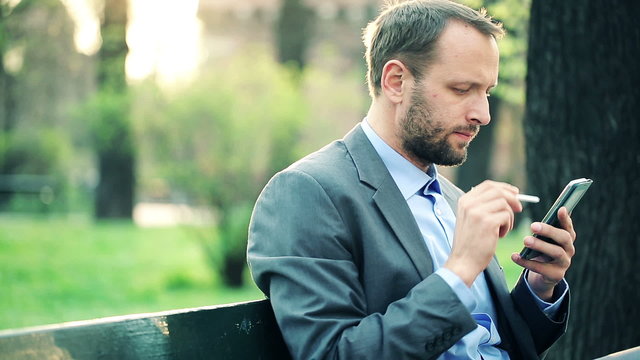 Young businessman with modern smartphone sitting on bench in par