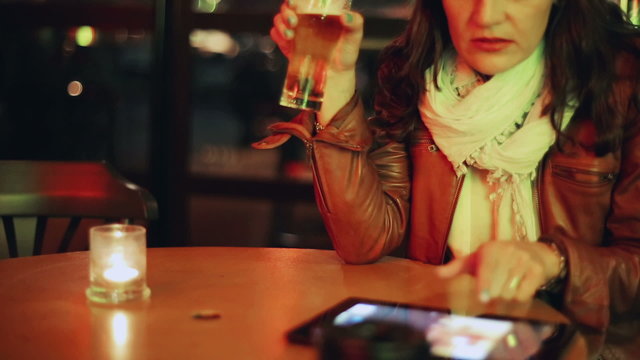Woman drinking beer and watching photos on tablet in pub, steady