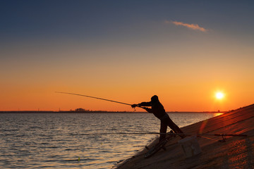 Silhouette of fisherman fishes on river bank against a sunset