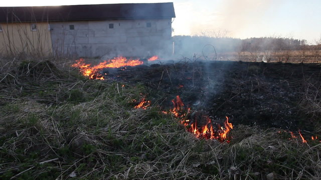 Fire in dry uncultivated grass near farm building during springtime