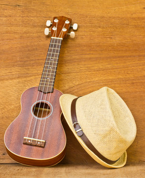 The ukulele and a hat is placed on a wooden floor.