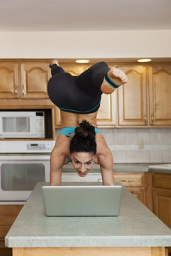 A woman doing a handstand balancing on a kitchen top and checking her laptop.