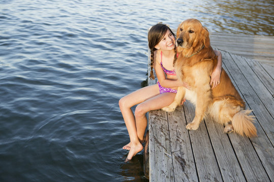 A girl and her golden retriever dog seated on a jetty by a lake.