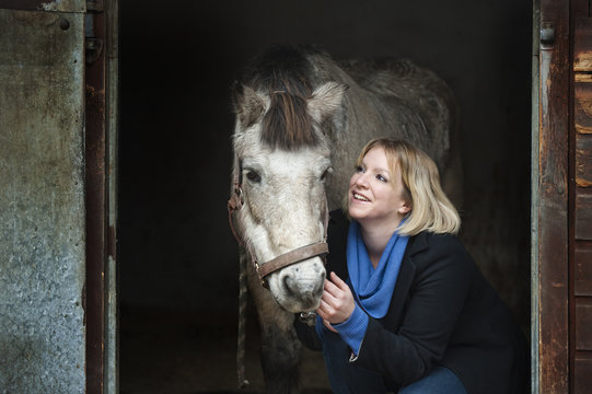 A woman standing next to a grey horse stroking its muzzle, at the stable door.