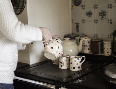 A woman pouring tea, by a range cooker. Using decorative china.