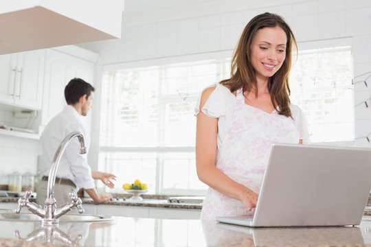 Woman Using Laptop With Man In Background In Kitchen