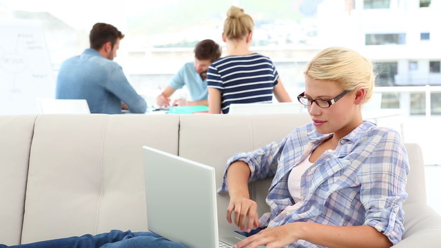 Blonde woman using her laptop on sofa with colleagues behind