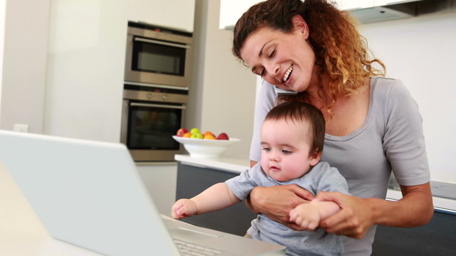Mother sitting with baby boy on lap using laptop and talking on