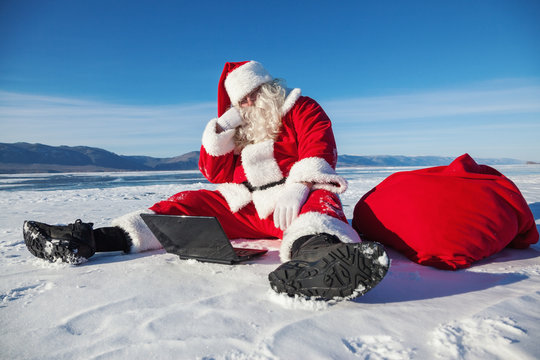 Santa Claus sitting on snow, looking at laptop news
