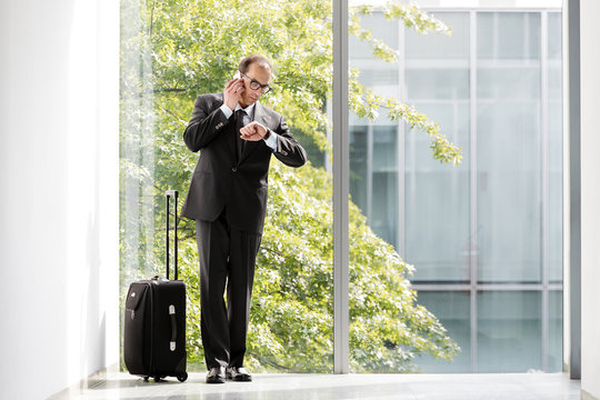 Businessman with trolley case looking at his watch and talking o