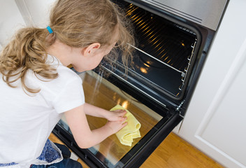 Little girl wipes oven in the kitchen at home.