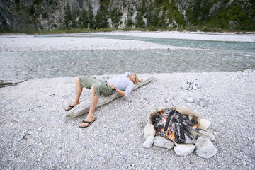 Obraz premium Deutschland,Bayern,Tölzer Land,Junger Mann auf Baumstamm in der Nähe von Lagerfeuer ruhen