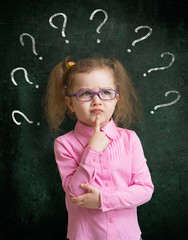 Child in eyeglasses standing near school blackboard with many qu