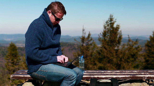 man working on a laptop and sitting on a bench in the mountainou
