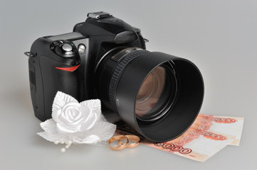 Photo camera, wedding boutonniere, rings with money on gray