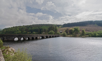 An old, stone bridge, with arches, spanning a tranquil lake