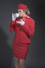 Retro blonde stewardess wearing red suit with cap. Drinking a cu