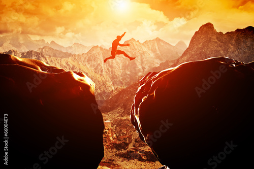 Man Jumping Over Precipice Between Two Mountains At Sunset