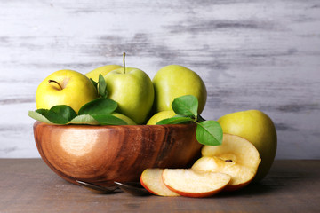 Ripe sweet apples with leaves in bowl on wooden background
