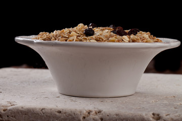 Side view of bowl of homemade granola with oats, almonds, organic raisins, and walnuts on a black background and travertine tile