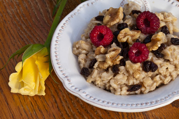 Top view of a bowl of oatmeal with walnuts, raisins, and red raspberries next to a yellow rose on a weathered wooden chair