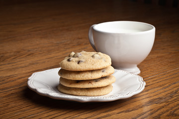 Stack of freshly baked chocolate chip cookies on a wooden table next to an antique teacup filled with milk