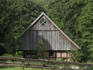 Weide mit Holzgatter und altem Bauernhaus aus Holz mit Spitzgiebel im Sommer am Waldrand im...