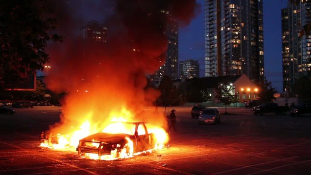 Two cars burn in parking lot with highrise at dusk