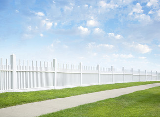 White fence, grass, sidewalk, blue sky and clouds