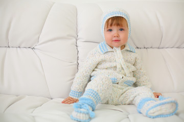 Cute little kid in knitted suit and hat sits on white couch