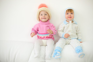 Two little kids in hats sit on back of white sofa at home.