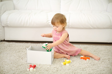 Little girl in dress puts toys in box near white sofa at home