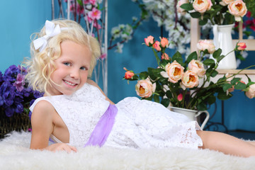 Blonde little girl in white dress lies on white fur and smiles