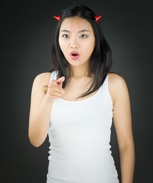 Shocked Asian young woman in devil horns pointing and pointing