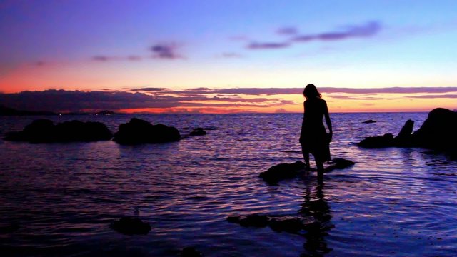 Silhouette of woman walking at a rocky beach, rises on stone