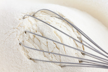 Whisk lying in white wheat flour in a glass dish.