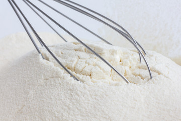 Whisk lying in white wheat flour in a glass dish.