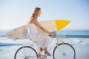 Beautiful surfer in sundress on bike holding surfboard at the be