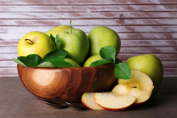 Ripe sweet apples with leaves in bowl on wooden background