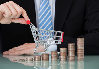 Businessman With Shopping Cart And Coin Stacks In Office