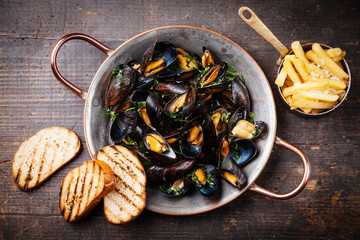Mussels in copper cooking dish and french fries on dark wooden b