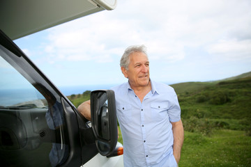 Senior man standing by camper, scenery in background