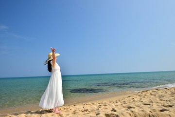 Fototapeta na wymiar 麦わら帽子と白いドレスを着てビーチに立っているアジア人女性の後ろ姿