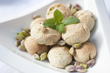 Handmade pistachio cookies with pistachios placed around and mint leaf placed on top in a white bowl on a white background.