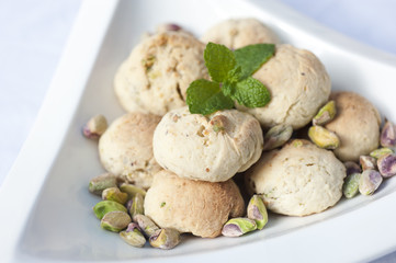 Handmade pistachio cookies with pistachios placed around and mint leaf placed on top in a white bowl on a white background.