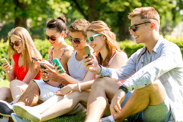 smiling friends with smartphones sitting on grass